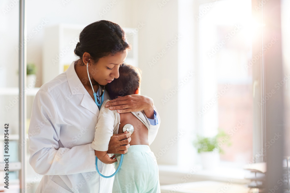 Wall mural Female doctor in white coat using stethoscope and listening to her little patient's heart at hospital