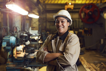 Waist up portrait of mixed-race female worker posing confidently while standing with arms crossed in factory workshop - obrazy, fototapety, plakaty