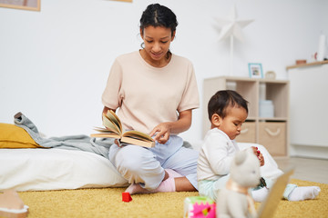 Young mother sitting on the floor and reading a book to her little child while he playing with toys