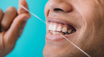 Close-up male mouth while flossing teeth with dental floss on blue background. Dental hygiene