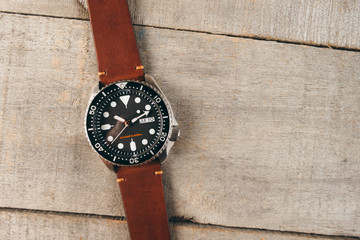 Men's luxury watch with black bezel, black dial and brown leather strap laying on the light wooden surface.