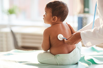 Rear view of shirtless toddler sitting on the table while doctor listening to him with stethoscope - obrazy, fototapety, plakaty
