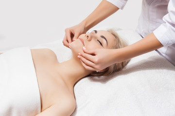 Concept of therapeutic anti-aging skin tightening facial treatments. Close-up hands of an unidentified young woman masseur doing facial massage to beautiful young caucasian client woman at spa salon.