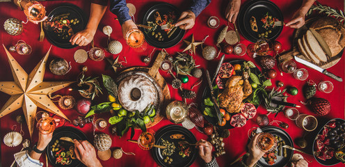 Friends celebrating Christmas. Flat-lay of people feasting over festive table with red cloth with champagne, roasted chicken, bundt cake, fruits and decorations, top view. Winter holiday party
