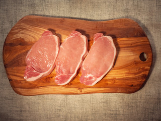 Three fresh uncooked raw port chops on a wooden cutting board. Simple white table cloth.