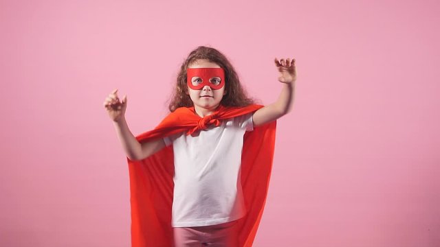 Beautiful little curly girl wearing red hero suit and mask at isolated over pink background.