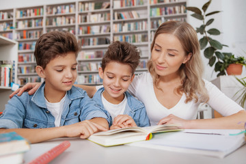 Fototapeta premium Beautiful woman helping her twin sons with homework, reading a book at the library. Two little brothers studying with their mom