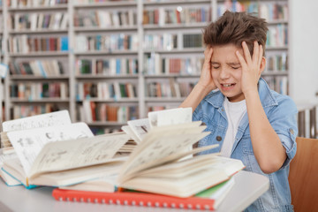 Young boy stressing out over pile of books and textbooks on his desk. Tired little student exhausted after studying