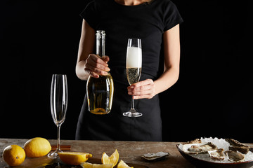 cropped view of woman holding glass and bottle with sparkling wine neae oysters in bowl with ice isolated on black