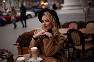 Beautiful european woman sit in the terrace of cafe in france style , holding a cup of latte with airy milk froth . The female enjoy the retro dtreet of the city. Model wearing coat and hat