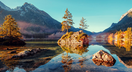 Amazing nature scene, trees on a rock island, glowing in sunlight, in Lake Hintersee at sunrise in...
