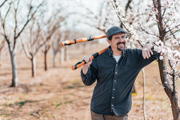 Positive farmer in blooming orchard with large pruning shears, leaning on a tree.