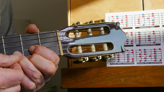 Close POV shot of a man learning to play classical guitar while sitting on a bed, with a hand carefully placing placing fingers on the fretboard, alongside a scale positioning chart. 	