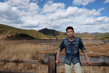 One Asian young man lean on handrail of wooden bridge. Withered reeds and mountain as background. Sunny blue sky with white clouds