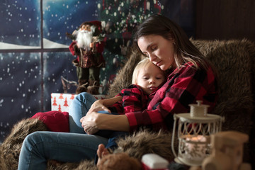Beautiful young mother, hugging her toddler boy, sitting in cozy chair on Christmas