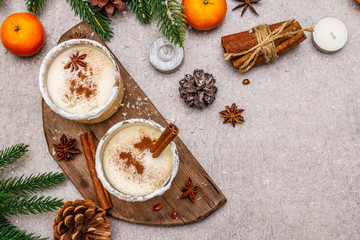 Obraz na płótnie Canvas Eggnog with cinnamon and nutmeg for Christmas and winter holidays. Homemade beverage in glasses with spicy rim. Tangerines, candles, gift. Stone concrete background