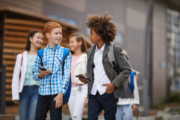 Two schoolboys using their mobile phones and talking to each other while walking along the street with other school children in the background