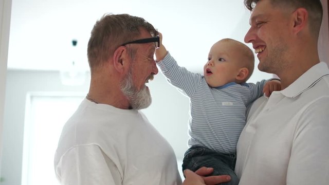 Toddler removes his glasses from his grandfather. Father holds the baby in his arms and laughs. Three generations of men of the same family, portrait of Indoor