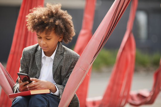African boy sitting on the swing with book and using his mobile phone on the playground outdoors