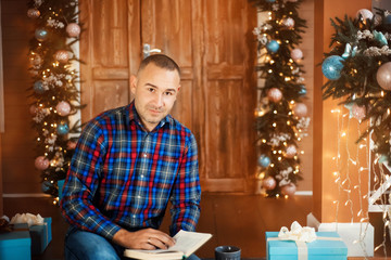 Man in plaid shirt with book reads on christmas decorated terrasse with gift boxes around. Male is looking in camera. Reading