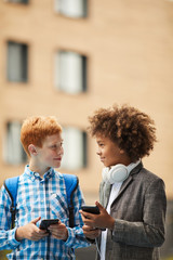 Two smiling boys standing and talking to each other and using their smart phones outdoors