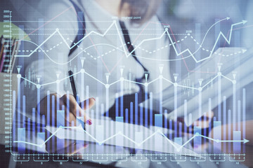 A woman hands writing information about stock market in notepad. Forex chart holograms in front. Concept of research.