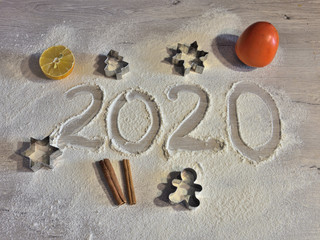 year 2020 written by hand on flour surface with Christmas fruits and spices cinnamon, orange, lemon and persimmon, and cookie cutters in shape of a man, a fir tree, a star and a snowflake