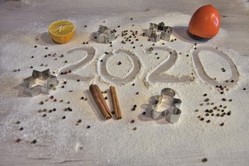 year 2020 written by hand on flour surface with Christmas fruits and spices cinnamon, pepper, orange, lemon and persimmon, and cookie cutters in shape of a man, a fir tree, a star and a snowflake