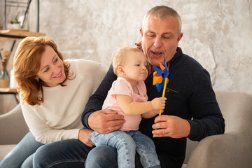 Happy family sitting on the sofa with sweet baby girl and blow on a windmill. Grandfather and grandmother spend their day together with granddaughter indoor