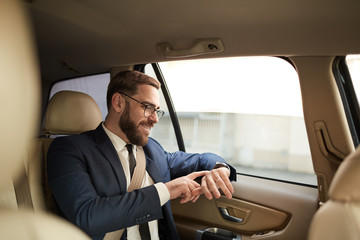 Happy young businessman in eyeglasses sitting on back seat and looking at his wristwatch while travelling by taxi