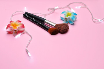 Obraz na płótnie Canvas Flat lay composition with two cosmetic makeup brushes, two hairpins in form of beautiful tropical flowers pink red and bright blue color supplemented with garland white lights on the pink background