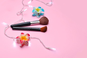 Obraz na płótnie Canvas Flat lay composition with two cosmetic makeup brushes, two hairpins in form of beautiful tropical flowers pink red and bright blue color supplemented with garland white lights on the pink background