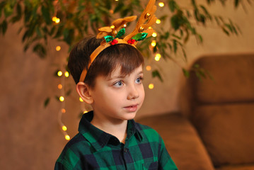 Cute little kid boy looks to the right wearing Christmas deer horns dressed up in green shirt with garland lights bokeh. Xmas winter holiday children lifestyle portrait. Beautiful New Year people