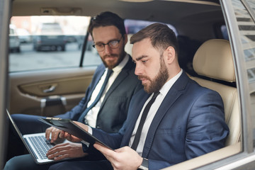 Two business partners using laptop and discussing document together on the back seat during driving