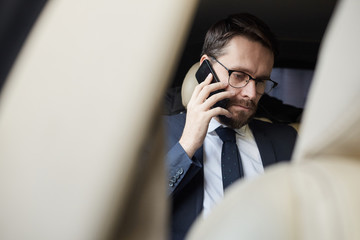 Young bearded businessman in eyeglasses sitting on back seat of the car and talking on mobile phone