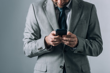 Cropped view of man in suit holding smartphone on grey background