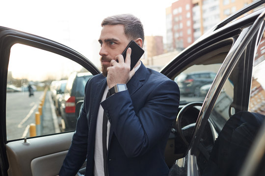 Young Bearded Businessman In Suit Coming Out Of The Car While Talking On Mobile Phone In The City