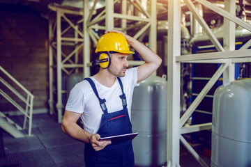 Worry handsome caucasian worker in overalls, with hardhat and antiphons holding tablet and standing in factory.