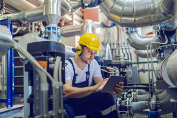 Serious caucasian handsome worker in overall, with hardhat and antiphons crouching and using...