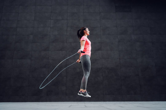 Side view of attractive fit caucasian brunette in sportswear and with ponytail skipping rope outdoors.