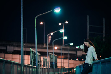 Plakat asian young woman with long hair in white top and jeans standing alone on the bridge at night looking at the ground and arm raised touching her hair feeling lonely and missing someone