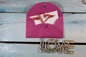 On a blue plank background, an open pink envelope from which a sheet of white paper with a red heart pierced by an arrow is visible. Below is the word "Love."