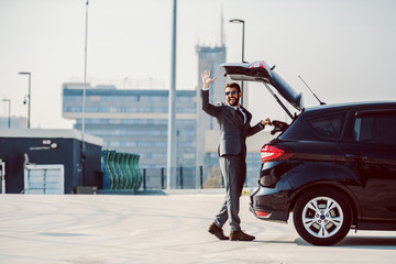 Full length of handsome caucasian bearded businessman with sunglasses taking his leather bag from trunk and waving.