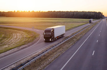 A caravan of modern freight trucks transports cargo in the evening against the sunset. Trucks drive...