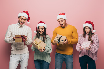 beautiful surprised friends in santa hats holding christmas presents, isolated on pink