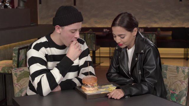 Couple eating fast food with burger and fries in modern stylish interior restaurant. Young man is feeding his lovely girlfriend with French fries. Happy moments together