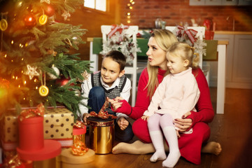 Fototapeta na wymiar Family - mother, son and daughter - playing and smiling on floor near christmas tree with gift boxes around. Woman, boy and girl - brother and sister - are happy and surprised in christmas time