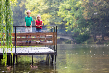 Two children boy and girl standing on wooden deck on a lake shore feeding ducks