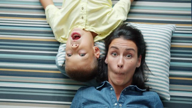 Portrait of mother and little boy son making funny faces lying on floor at home having fun together. Joyful people, happy family and positive emotions concept.