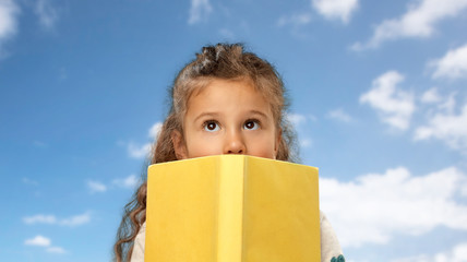 reading, education and childhood concept - little girl hiding behind yellow book over blue sky and clouds background